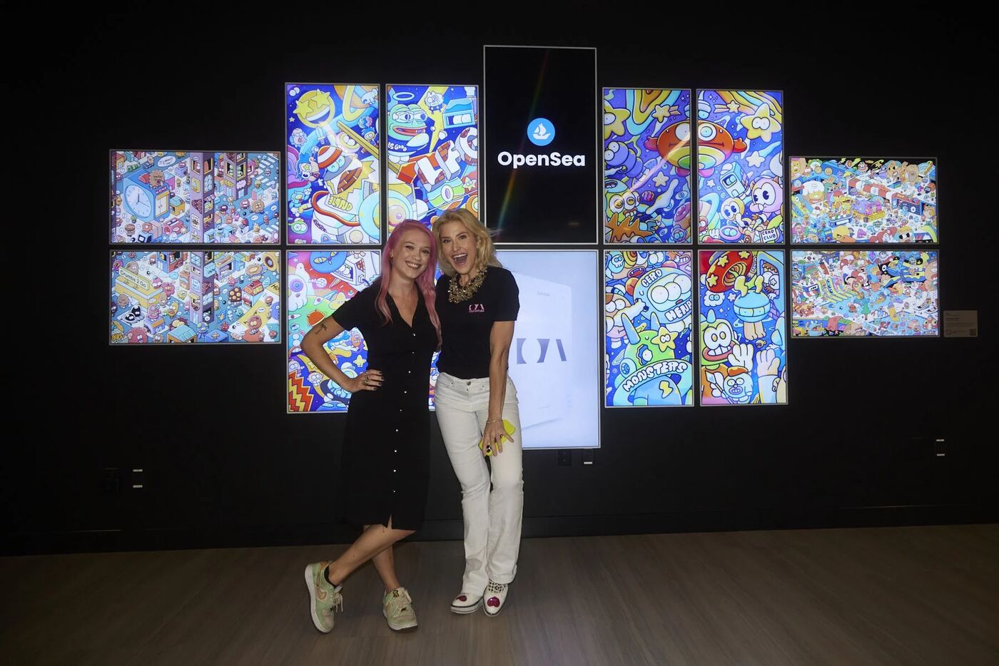 Two hosts posing in front of the OpenSea digital art wall